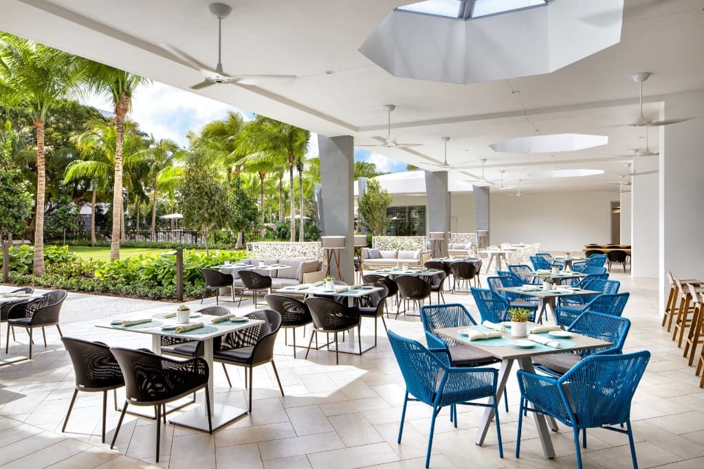 Covered terrace dining: white tables with teal placemats, woven chairs, ceiling fans, and palm trees on the lawn beyond