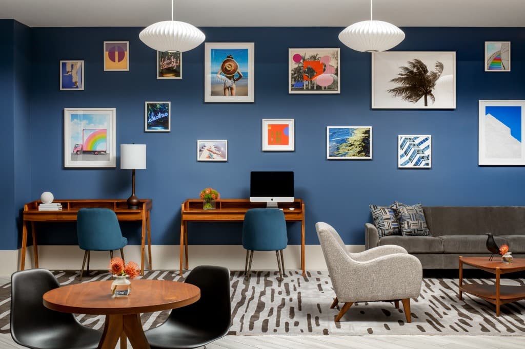 Business center lounge: blue gallery wall with framed art, wood writing desks, teal chairs, and a grey sofa on a patterned rug