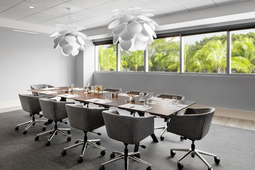 Seven Isles boardroom: long wood conference table with grey chairs, sculptural pendant lights, and palm trees through the window