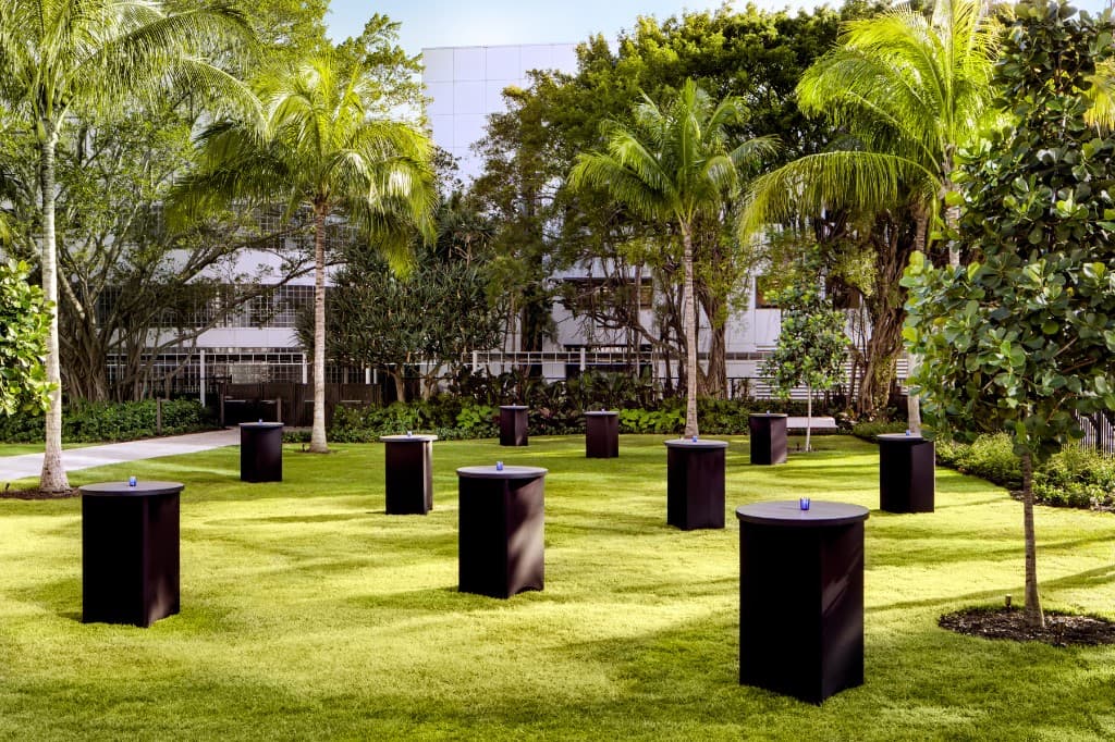 West Lawn cocktail setup with high tables on green lawn, tropical palms, and hotel building beyond