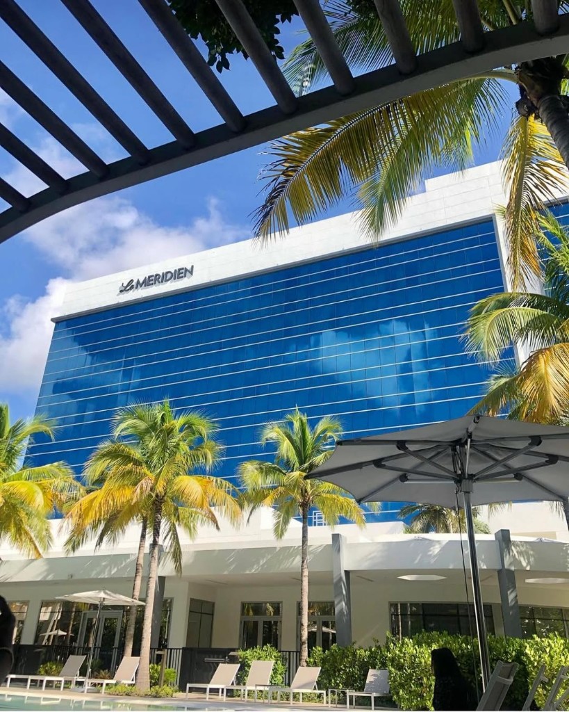 Resort pool deck with loungers, palm trees, and glass tower under a bright sky